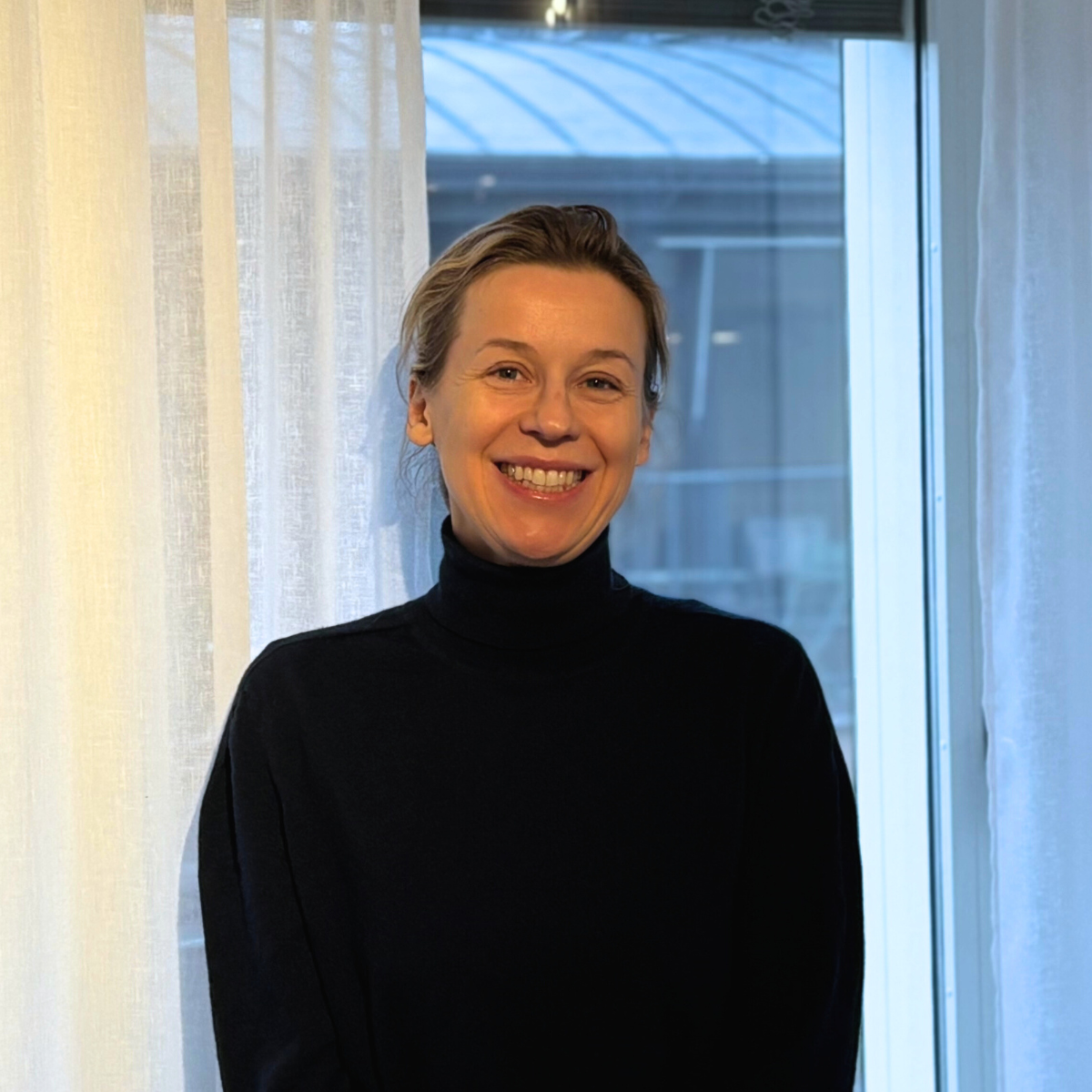 Person i svart tröja står framför ett fönster med ljusa gardiner, i en inomhusmiljö.