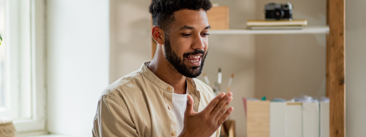 En person i beige skjorta gestikulerar med händerna i ett ljust rum med hyllor och en kamera i bakgrunden.